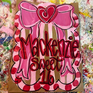 Op de afbeelding: Een bruine papieren cadeauzak met een roze strik en hartontwerp. De tas heeft verticale strepen en de woorden "Mackenzie Sweet 16" in rood schrift. De strik is zwart omlijnd.