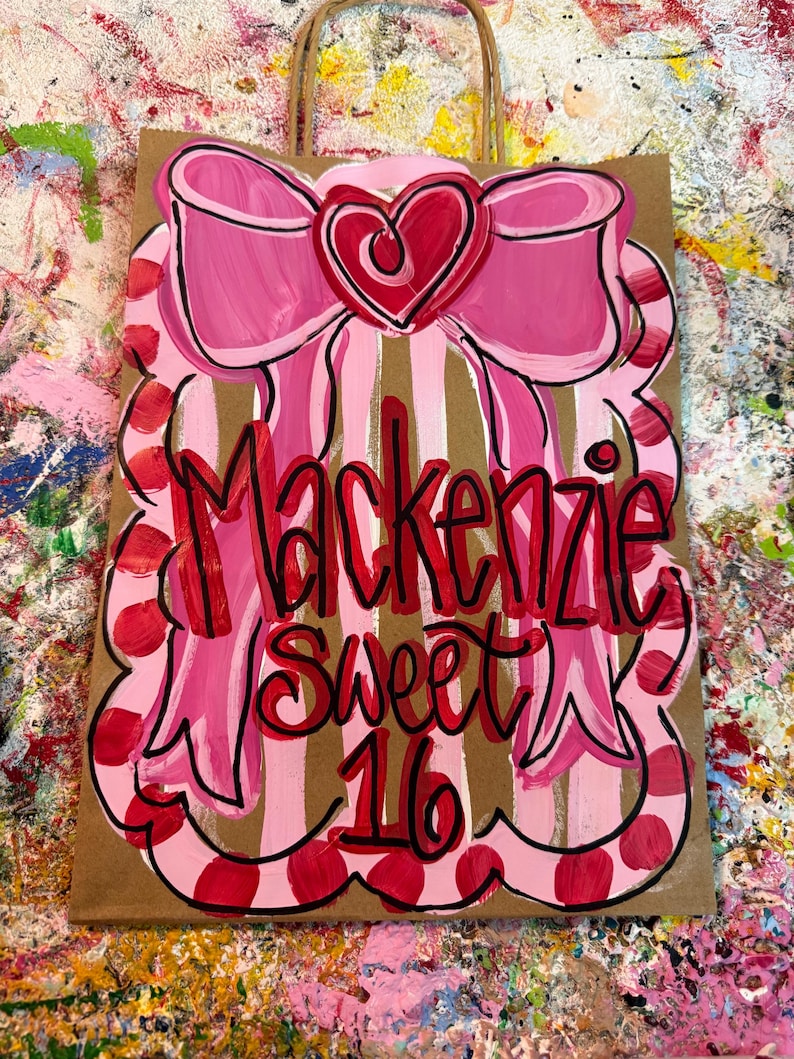 Op de afbeelding: Een bruine papieren cadeauzak met een roze strik en hartontwerp. De tas heeft verticale strepen en de woorden "Mackenzie Sweet 16" in rood schrift. De strik is zwart omlijnd.