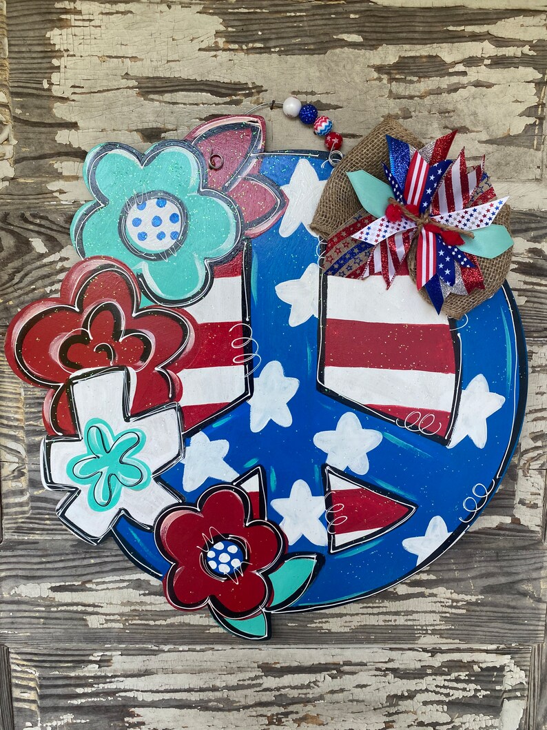 May include: A blue and red peace sign with white stars and red stripes, decorated with red, white, and blue flowers. The peace sign is made of wood and has a rustic look.