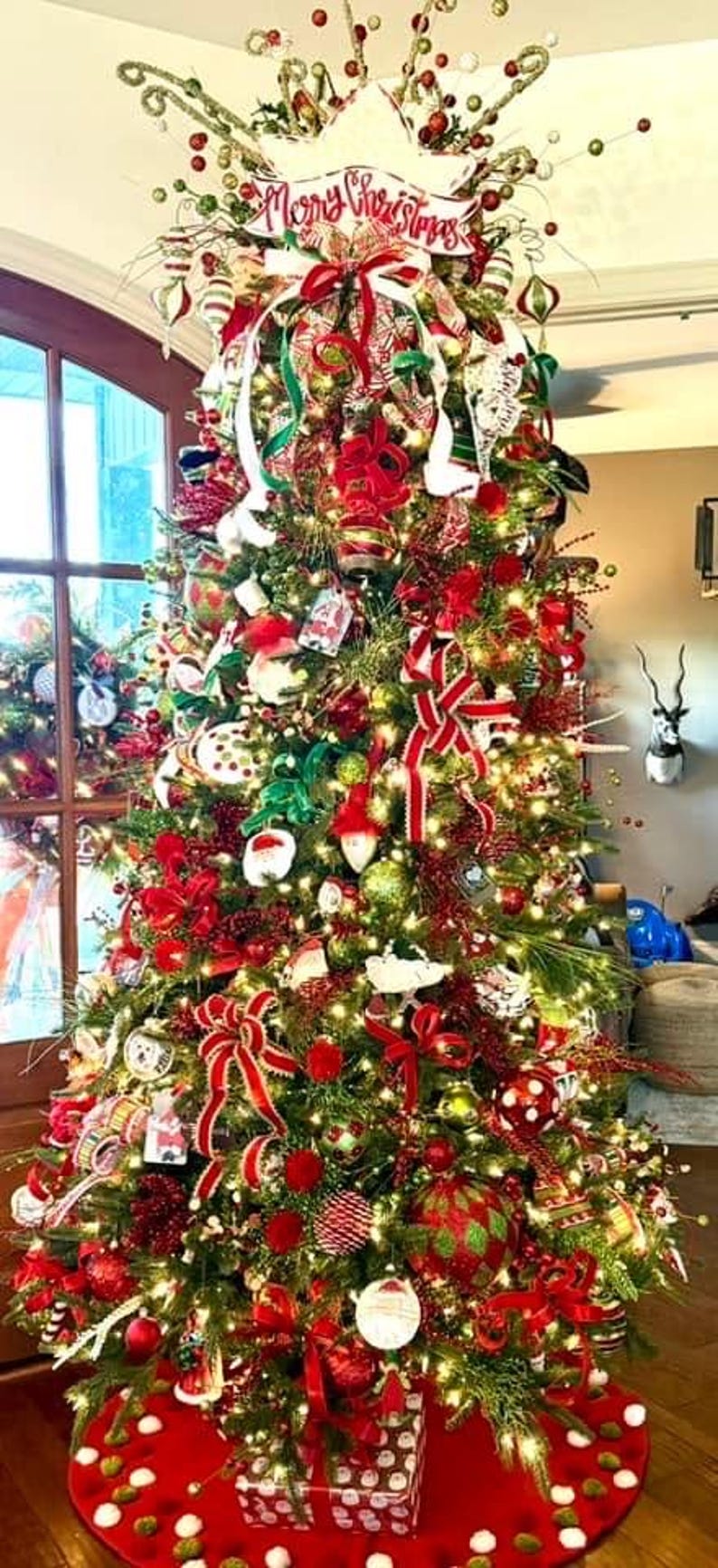 May include: A decorated Christmas tree with red, green, and white decorations, bows, and lights. The tree is topped with a sign saying "Merry Christmas". A red tree skirt and wrapped gifts are at the base.