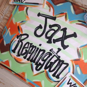 May include: A colorful wooden sign with the name "Jax Remington" written in black paint. The sign has a green, orange, and blue abstract design. There are also two smaller white signs with the words "Time:" and "Weight lbs." written in black.