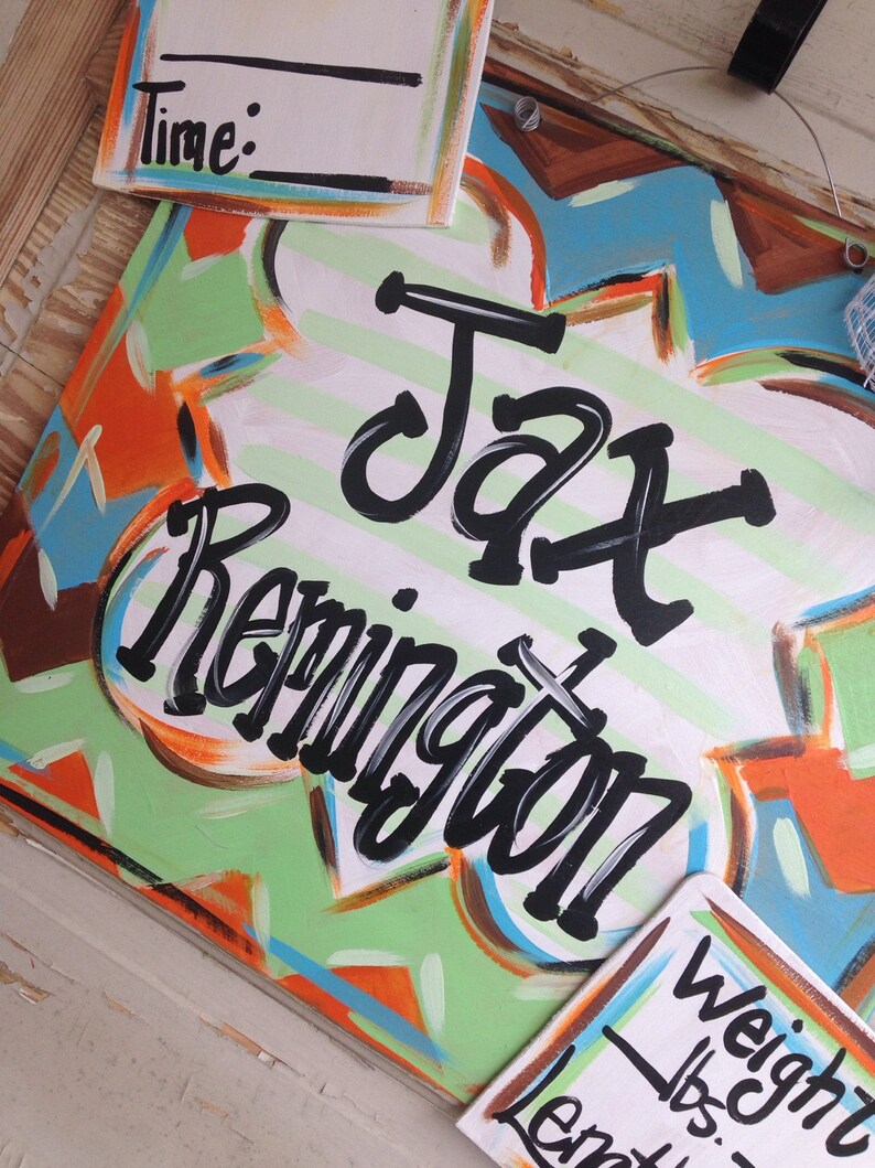 May include: A colorful wooden sign with the name "Jax Remington" written in black paint. The sign has a green, orange, and blue abstract design. There are also two smaller white signs with the words "Time:" and "Weight lbs." written in black.