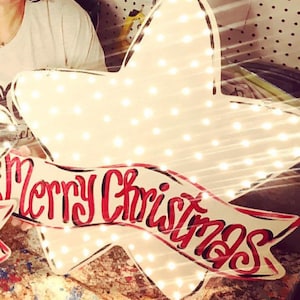 May include: A large, illuminated star-shaped Christmas decoration. The star is white with many small lights. A banner across the star reads "Merry Christmas" in red script. Suitable for festive indoor or outdoor display.