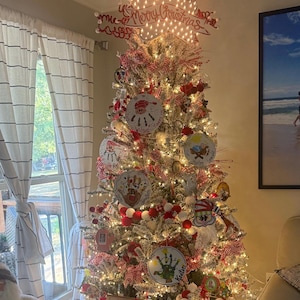 May include: A decorated Christmas tree with white lights, red and white ribbons, and ornaments. The tree topper reads "Merry Christmas". Ornaments include family photos and festive designs. A garland of red and white pom-poms adorns the tree.