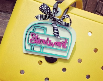 May include: A white camper van charm with the word "Stoodvant" in pink letters. The charm is attached to a black and white polka dot ribbon and a black carabiner clip.
