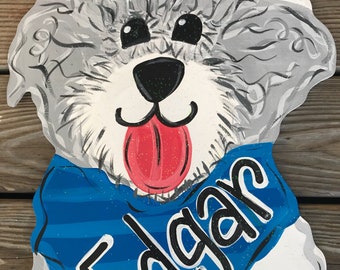 May include: A hand-painted wooden door hanger shaped like a dog with gray and white fur, black eyes, a red tongue, and a blue bandana with the name "Edgar" written in white.