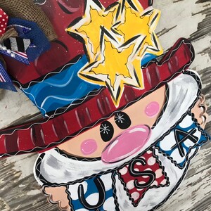 May include: A wooden door hanger featuring a cartoon Uncle Sam with a red, white, and blue hat, a white beard, and a pink nose. The hat has two yellow stars on it. The hanger has a banner that says "USA" in red, white, and blue.