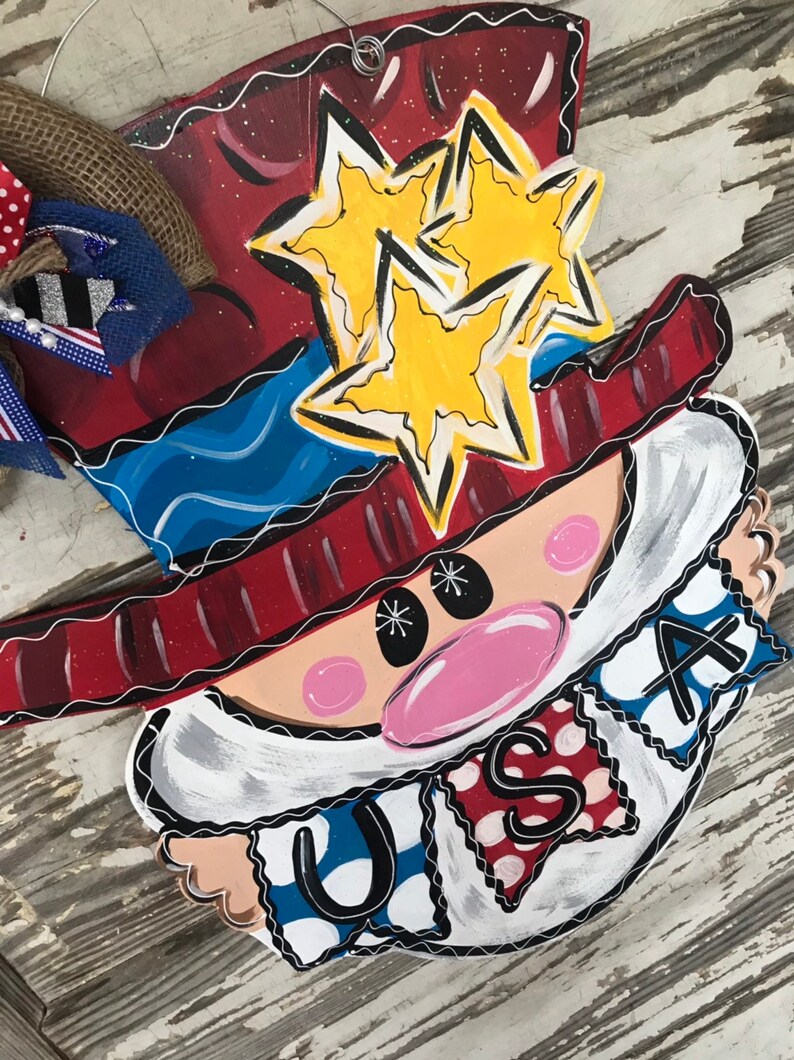 May include: A wooden door hanger featuring a cartoon Uncle Sam with a red, white, and blue hat, a white beard, and a pink nose. The hat has two yellow stars on it. The hanger has a banner that says "USA" in red, white, and blue.