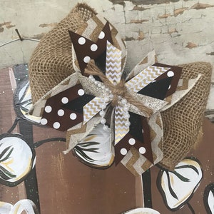 May include: A brown, white, and gold bow with a burlap base. The bow is decorated with white polka dots, gold glitter, and white beads. The bow is tied with a brown twine.
