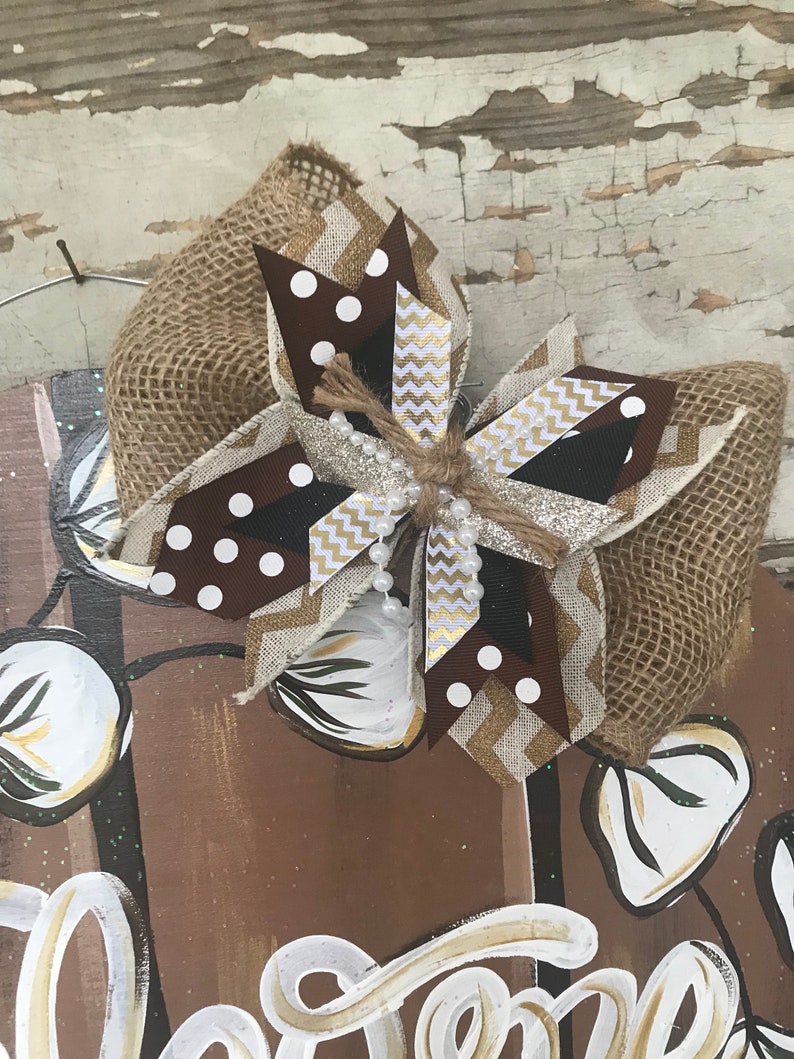 May include: A brown, white, and gold bow with a burlap base. The bow is decorated with white polka dots, gold glitter, and white beads. The bow is tied with a brown twine.