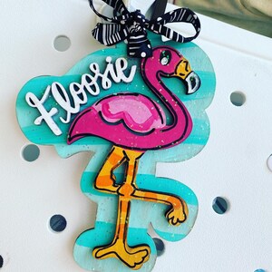 May include: A pink flamingo with glitter accents stands on a turquoise and white striped background. The flamingo has a black eye and a yellow beak. The word "Floosie" is written in white on the background.