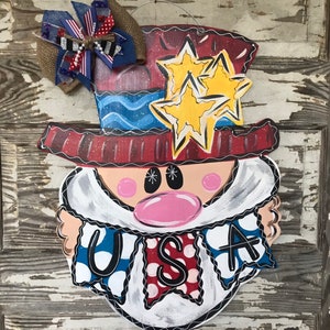 May include: A wooden door hanger shaped like a cartoon face wearing a red, white, and blue hat with stars. The face has a pink nose and white beard. The face is holding a banner that says "USA" in black letters.
