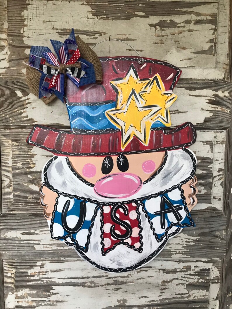 May include: A wooden door hanger shaped like a cartoon face wearing a red, white, and blue hat with stars. The face has a pink nose and white beard. The face is holding a banner that says "USA" in black letters.