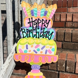 Könnte beinhalten: Bunte Holzdekoration für einen Geburtstagskuchen mit der Aufschrift "Happy Birthday". Der Kuchen hat mehrere Schichten, Kerzen und dekorative Streusel. Er ist in leuchtenden Pink-, Gelb-, Grün- und Orangetönen bemalt.