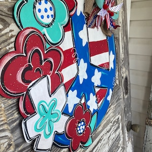 May include: A wooden peace sign door hanger decorated with red, white, and blue flowers and stars. The peace sign is painted in blue and white with red and white stripes. The flowers are painted in red, white, and blue with white polka dots. The door hanger is decorated with a red, white, and blue bow.