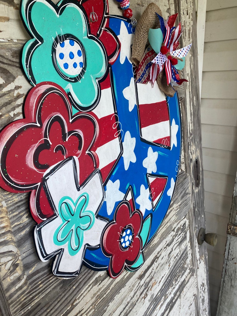May include: A wooden peace sign door hanger decorated with red, white, and blue flowers and stars. The peace sign is painted in blue and white with red and white stripes. The flowers are painted in red, white, and blue with white polka dots. The door hanger is decorated with a red, white, and blue bow.
