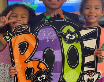 May include: Three children hold a large wooden BOO sign decorated with Halloween themes. The sign is painted in orange, purple, green, and black with a spider, bat, and heart accents.