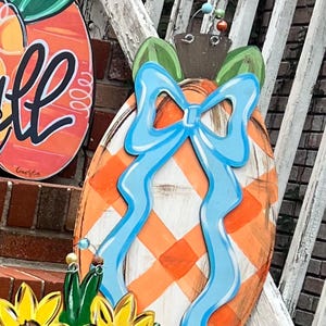 Puede incluir: Un colgador de puerta de calabaza de madera con un lazo azul y un patrón de cuadros blancos y naranjas. La calabaza está decorada con hojas verdes y se apoya en una barandilla de madera blanca.