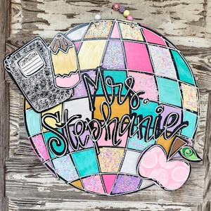 May include: A colorful globe with a pink, yellow, blue, and green checkered pattern. The globe has glitter accents and the text "Mrs. Stephanie" written in black. There is a black and white composition notebook, a yellow pencil, and a pink apple with a green leaf on the globe.