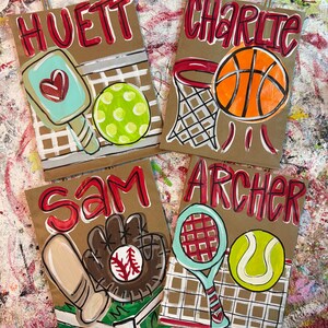 Op de afbeelding: Vier bruine papieren cadeauzakken met sportthema-illustraties. E&eacute;n tas toont een pickleball-peddel en -bal, een andere een basketbal en -mand, een derde een honkbal en -handschoen, en de laatste een tennisracket en -bal. Elke tas heeft een naam in het rood.