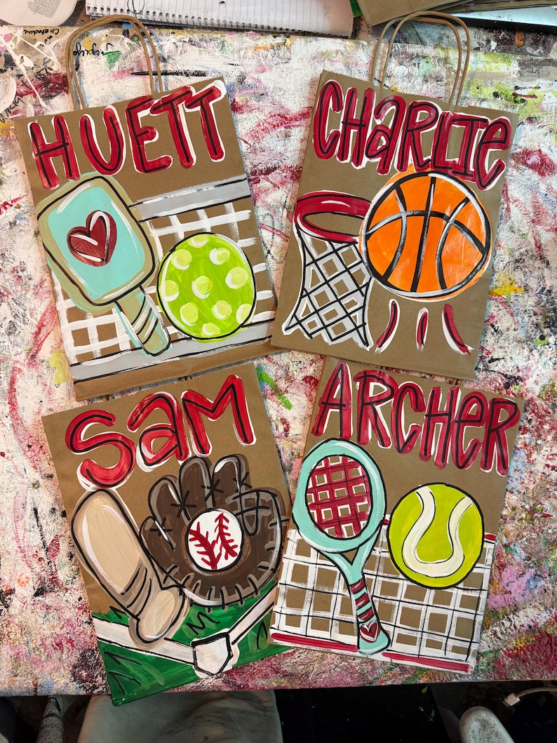 Op de afbeelding: Vier bruine papieren cadeauzakken met sportthema-illustraties. E&eacute;n tas toont een pickleball-peddel en -bal, een andere een basketbal en -mand, een derde een honkbal en -handschoen, en de laatste een tennisracket en -bal. Elke tas heeft een naam in het rood.