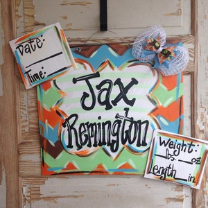 May include: A wooden sign with a chevron pattern in orange, green, and blue. The sign says "Jax Remington" in black cursive lettering. There are two white cards with black text for recording the date, time, weight in pounds and ounces, and length in inches.