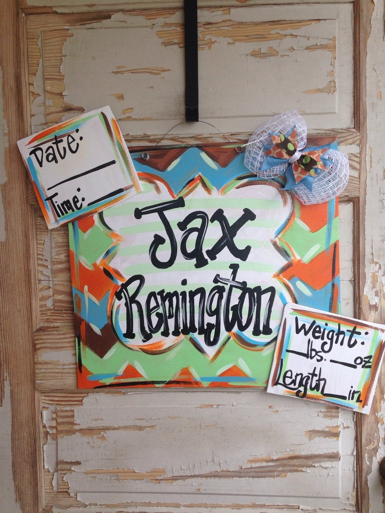 May include: A wooden sign with a chevron pattern in orange, green, and blue. The sign says "Jax Remington" in black cursive lettering. There are two white cards with black text for recording the date, time, weight in pounds and ounces, and length in inches.