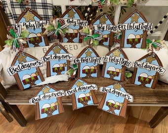 Peut inclure: Ornements de Noël en bois en forme d'étable, peints avec un fond de ciel bleu et une scène de crèche. Certains ont le texte "Oh Holy Night" ou "Best day ever" sur une bannière. Chaque ornement a une étoile dorée.