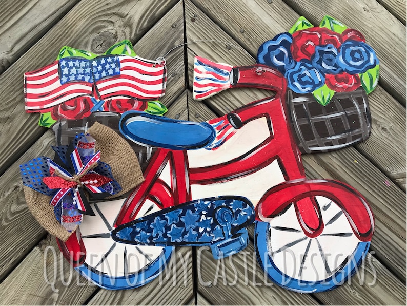 May include: A red, white, and blue tricycle with a basket of flowers on the handlebars. The tricycle has a blue and white star pattern on the wheels. The basket is filled with red, white, and blue flowers. The tricycle is on a wooden background.