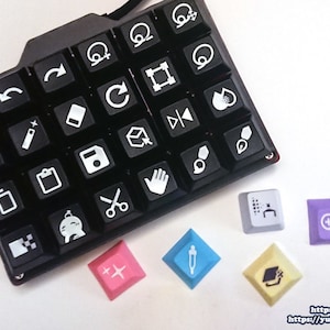Puede incluir: Un teclado negro con iconos blancos para diversas funciones de dise&ntilde;o y edici&oacute;n. El teclado tiene un dise&ntilde;o &uacute;nico con una forma curva. Hay seis teclas adicionales con diferentes iconos y colores, incluyendo una tecla azul con un icono de cuentagotas, una tecla amarilla con un icono de gorro de graduaci&oacute;n, una tecla morada con un icono de lupa, una tecla blanca con un icono de cuadr&iacute;cula y una tecla rosa con un icono de signo m&aacute;s.