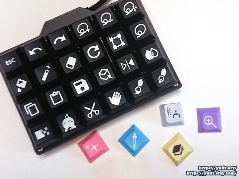 Puede incluir: Un teclado negro con iconos blancos para diversas funciones de dise&ntilde;o y edici&oacute;n. El teclado tiene un dise&ntilde;o &uacute;nico con una forma curva. Hay seis teclas adicionales con diferentes iconos y colores, incluyendo una tecla azul con un icono de cuentagotas, una tecla amarilla con un icono de gorro de graduaci&oacute;n, una tecla morada con un icono de lupa, una tecla blanca con un icono de cuadr&iacute;cula y una tecla rosa con un icono de signo m&aacute;s.