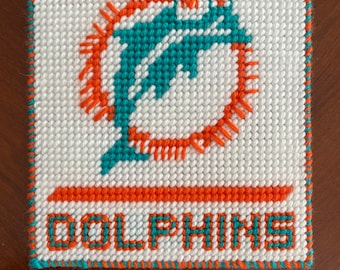 Puede incluir: Cuadrado de punto de aguja blanco con el logotipo de los Miami Dolphins. El logotipo incluye un delfín turquesa saltando a través de un sol naranja. La palabra "DOLPHINS" está cosida en naranja y turquesa. Los bordes están ribeteados en turquesa y naranja.