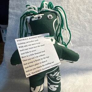 May include: A handmade green and white doll with googly eyes and yarn hair. The doll is holding a paper with the text "FOOTBALL DAMMIT DOLL" and a poem about football. The doll's legs feature a team logo.