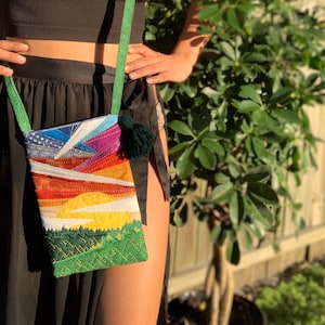 May include: A colorful patchwork fabric crossbody bag with a green tassel. The bag features a sunset scene with a yellow sun, orange and red clouds, and a green forest at the bottom.
