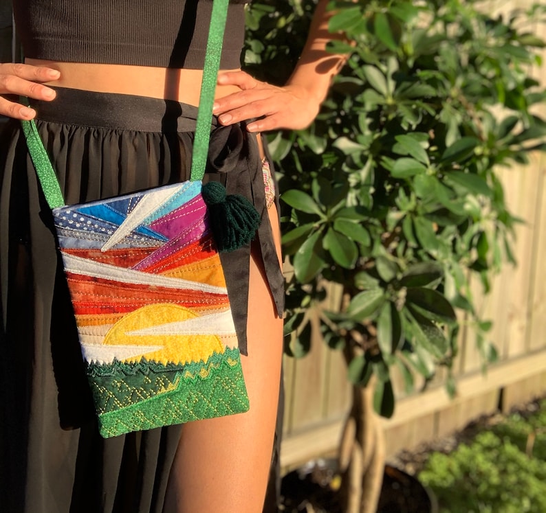 Peut inclure: Un sac &agrave; bandouli&egrave;re en tissu patchwork color&eacute; avec un pompon vert. Le sac repr&eacute;sente un coucher de soleil avec un soleil jaune, des nuages orange et rouges et une for&ecirc;t verte en bas.
