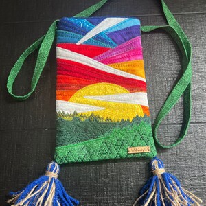 Peut inclure: Une pochette de t&eacute;l&eacute;phone matelass&eacute;e avec un motif de coucher de soleil color&eacute; et des montagnes vertes. La pochette a une sangle verte avec des glands bleus et une &eacute;tiquette brune qui dit "Life Made by Lu".