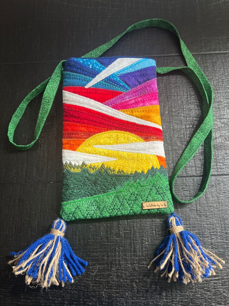 Peut inclure: Une pochette de t&eacute;l&eacute;phone matelass&eacute;e avec un motif de coucher de soleil color&eacute; et des montagnes vertes. La pochette a une sangle verte avec des glands bleus et une &eacute;tiquette brune qui dit "Life Made by Lu".