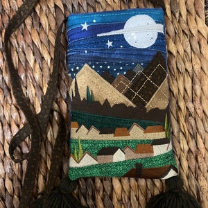 Peut inclure: Un petit sac matelass&eacute; avec une bandouli&egrave;re et des glands marrons. Le sac pr&eacute;sente un motif de ciel nocturne avec des &eacute;toiles, une lune et des montagnes. Le bas du sac a un paysage vert et marron avec des maisons.