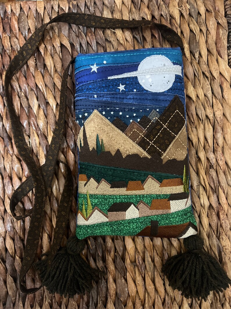 Peut inclure: Un petit sac matelass&eacute; avec une bandouli&egrave;re et des glands marrons. Le sac pr&eacute;sente un motif de ciel nocturne avec des &eacute;toiles, une lune et des montagnes. Le bas du sac a un paysage vert et marron avec des maisons.