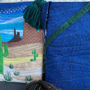 Peut inclure: Un sac en tissu matelass&eacute; avec une sc&egrave;ne de d&eacute;sert d'un c&ocirc;t&eacute; et un tissu &agrave; motifs bleus et verts de l'autre. La sc&egrave;ne du d&eacute;sert pr&eacute;sente un cactus, une montagne et un ciel bleu. Le sac a un pompon vert.
