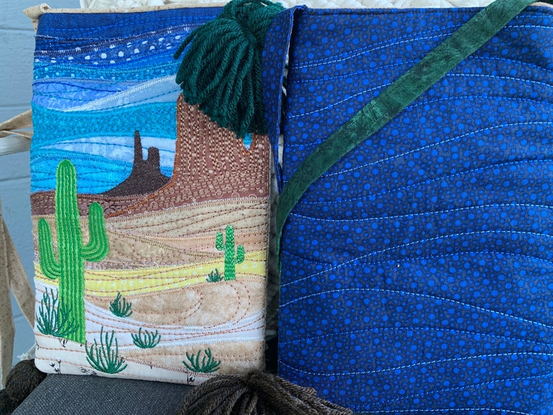 Peut inclure: Un sac en tissu matelass&eacute; avec une sc&egrave;ne de d&eacute;sert d'un c&ocirc;t&eacute; et un tissu &agrave; motifs bleus et verts de l'autre. La sc&egrave;ne du d&eacute;sert pr&eacute;sente un cactus, une montagne et un ciel bleu. Le sac a un pompon vert.