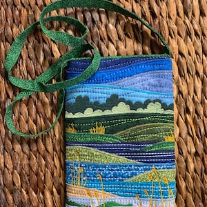May include: A small quilted bag with a green strap. The bag features a landscape scene with blue water, green hills, and yellow grass.