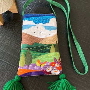 May include: A small quilted bag with a green strap and tassels. The bag features a colourful embroidered landscape with a blue sky, white clouds, green hills, and flowers.