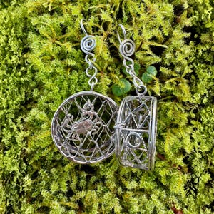 Könnte beinhalten: Silberne Ohrringe mit einem Krabbenmotiv in einem kreisförmigen, netzartigen Rahmen. Die Ohrringe hängen an einem Spiral- und Kettendesign. Der Hintergrund ist ein Bett aus leuchtend grünem Moos.