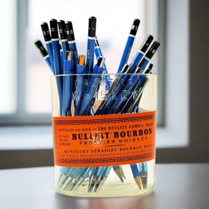 May include: A clear glass with a Bulleit Bourbon label filled with blue pencils and pens. The pencils have black tips and markings like "HB" and "2H". The pens are blue with silver accents. The glass sits on a light-colored surface.
