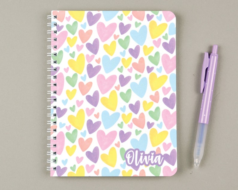 May include: A white spiral notebook with a colorful heart pattern and the name "Olivia" printed on the cover. The notebook is next to a purple pen.