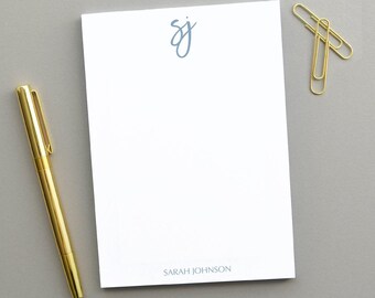 May include: A white notepad with a blue handwritten monogram "SJ" at the top and the name "SARAH JOHNSON" printed at the bottom. A gold pen and two gold paper clips are on a grey surface.
