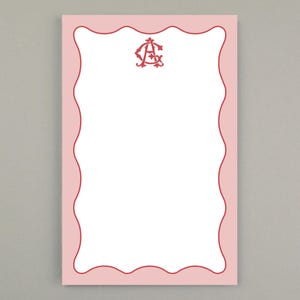 Puede incluir: Un bloc de notas rosa con un borde rojo ondulado y un monograma rojo en la parte superior. El bloc de notas tiene un interior blanco para escribir. El diseño es simple y elegante, adecuado para uso personal o profesional.