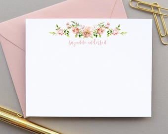 May include: White stationery card with a floral design in shades of pink and green. The card has the name "Suzanne Anderson" printed in a delicate pink script. A matching pink envelope is visible in the background, along with a gold pen and paperclip.