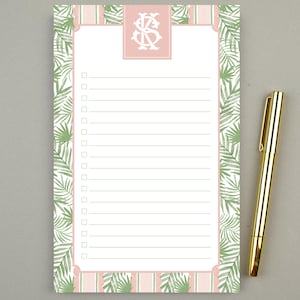 May include: A lined notepad with a pink and green tropical leaf design and a gold pen. The notepad features a pink border with a monogram and a checklist format. The pen is gold and rests on a gray surface.